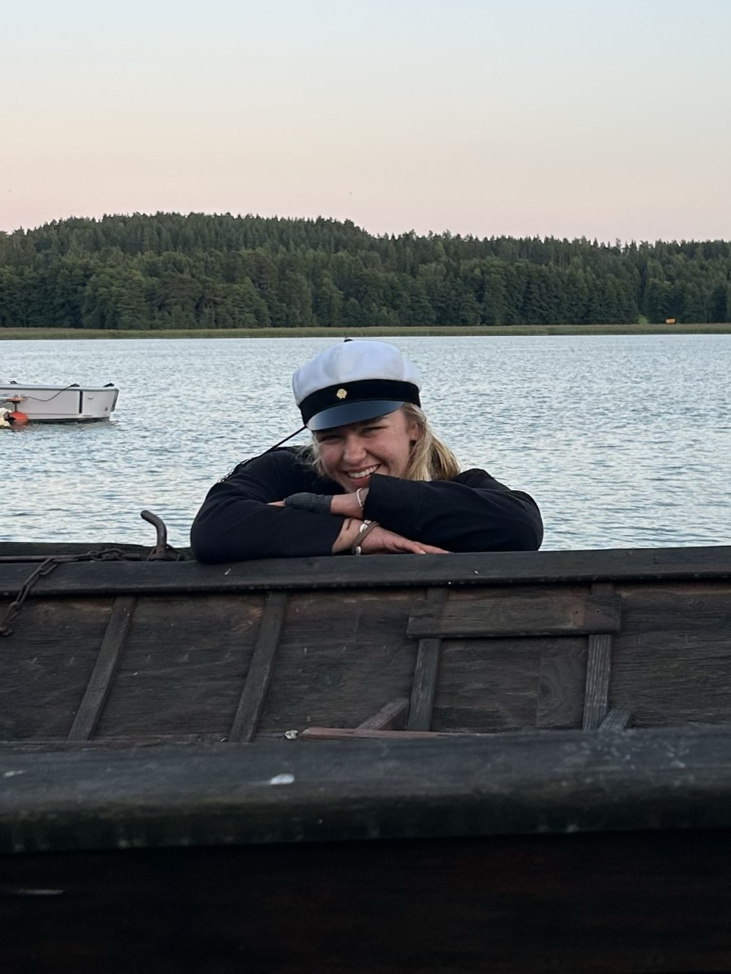 Leaning on the side of a wooden boat at dusk, wearing a student cap.